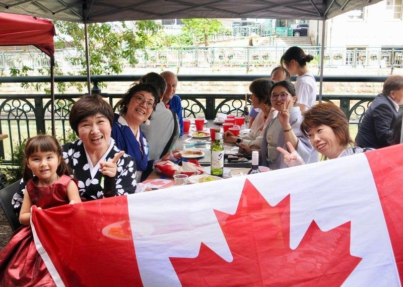 People at a table with a Canadian flag, smiling and gesturing; outdoor setting with greenery.