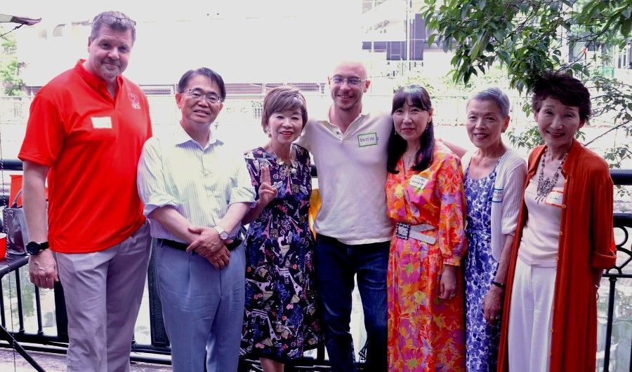 Group of people posing outdoors, smiling. Includes people in red, white, and floral clothing. Wooden railing and foliage in the background.