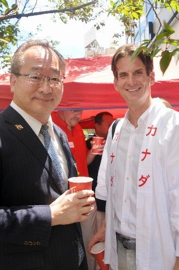 Two men smiling, holding red cups, in front of a red tent, one wearing a suit, the other a white shirt with Japanese writing.
