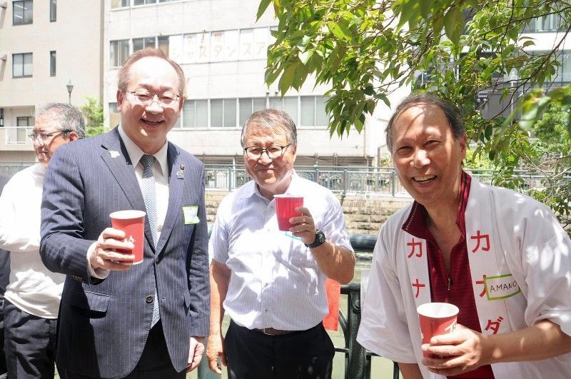 Three men smiling, toasting with red cups outdoors. Buildings and greenery in the background.