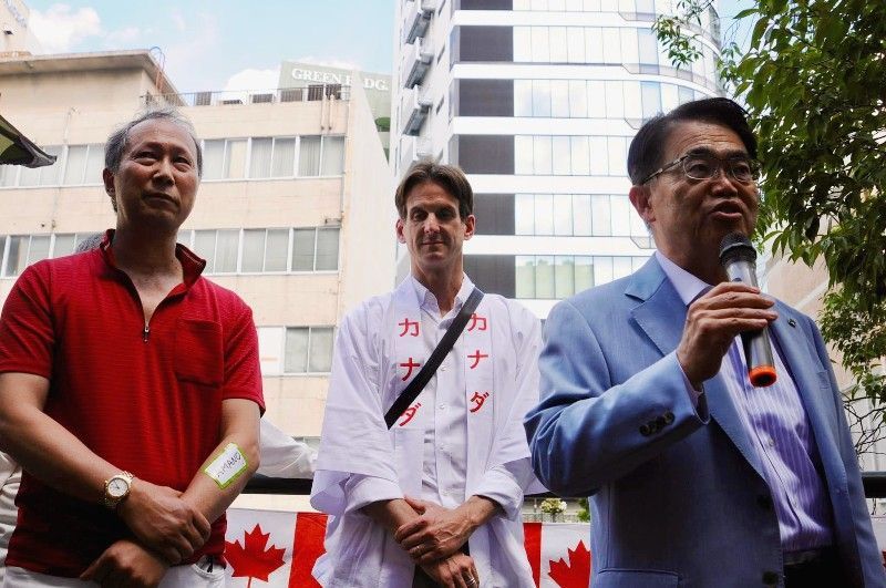 Three men standing outside; one speaks into a microphone, another wears a shirt with Japanese writing.