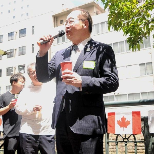 Man in suit speaks into microphone, holding a cup, near Canadian flags.