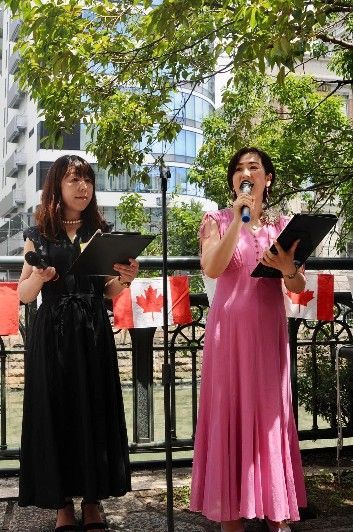 Two women speaking at an outdoor event with Canadian flags. One wears a black dress, the other a pink gown.