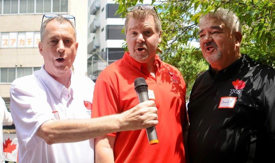 Three men singing outdoors, holding microphone, wearing red and white shirts.