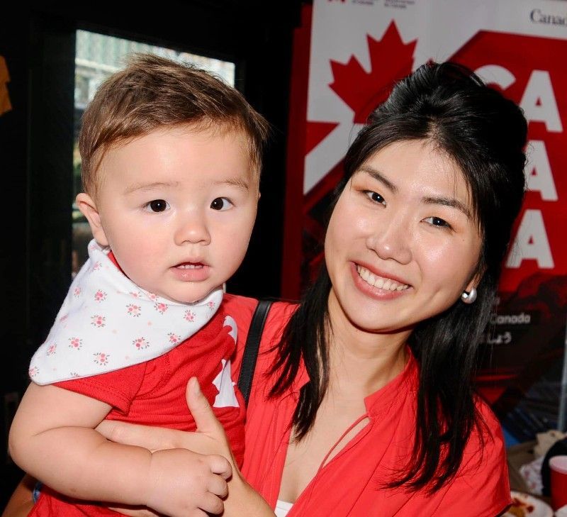Woman smiling, holding baby in red, both in front of a Canadian flag.
