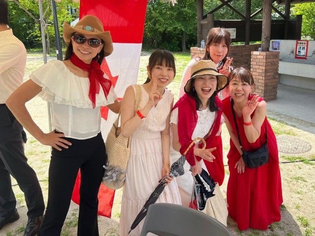 Five people smile, pose with Canadian flag; woman in cowboy hat. Outdoor setting, bright colors.
