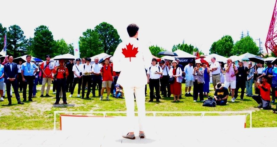 Man in white suit with Canadian flag on back, facing crowd outdoors.