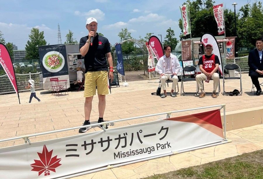 Man speaking at an event in Mississauga Park, Japan. Banner in Japanese and English, people sitting behind.