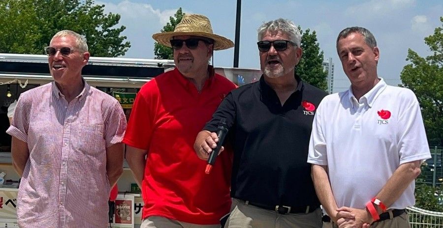 Four men standing outdoors, one holding a microphone. They are wearing casual shirts.