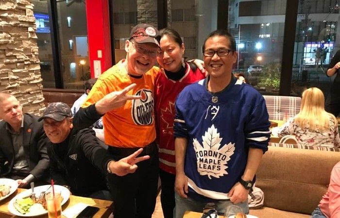 Three people smiling, posing for photo in a restaurant; one in Oilers shirt, one in Flames shirt, one in Maple Leafs jersey.