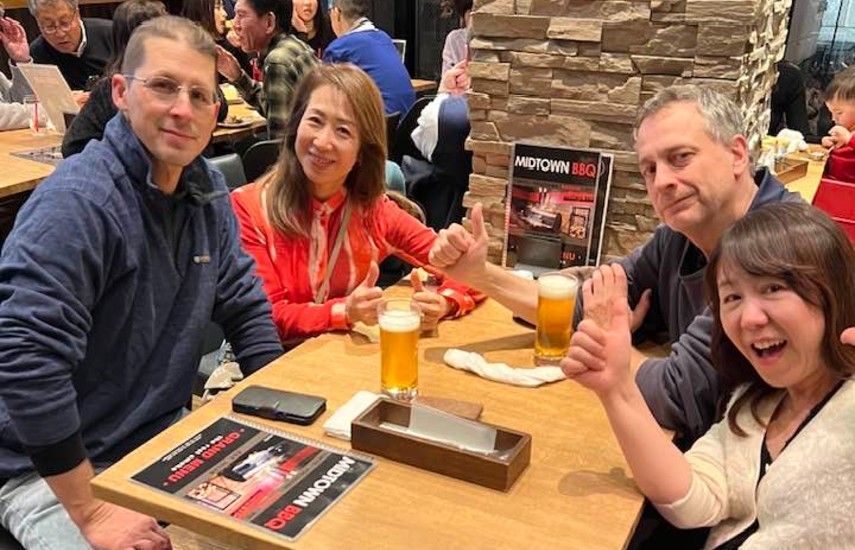 Four people at a table in a restaurant give thumbs up, holding drinks. Smiling and looking at camera.