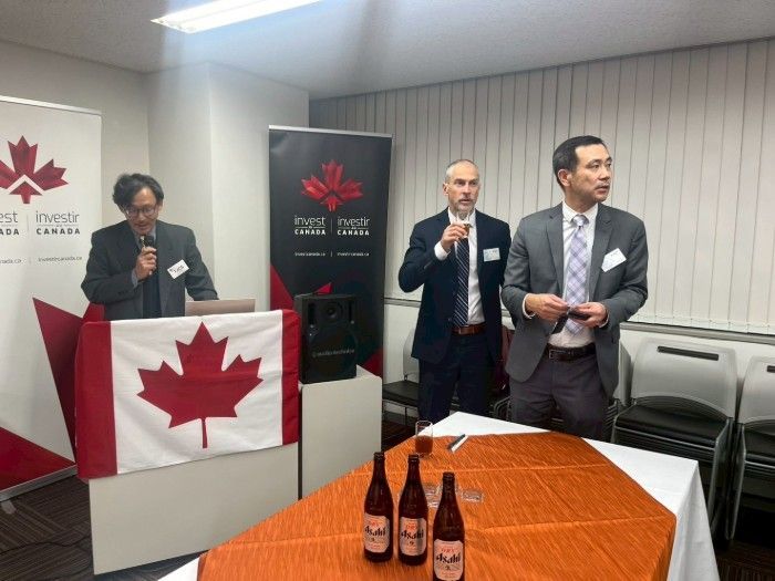 Three men at a speaking event in front of a Canadian flag and promotional banner; bottles of beer on a table.