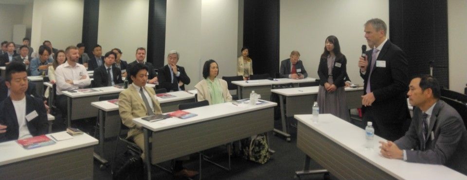 A conference room with a speaker addressing an audience seated at tables. People listen attentively.