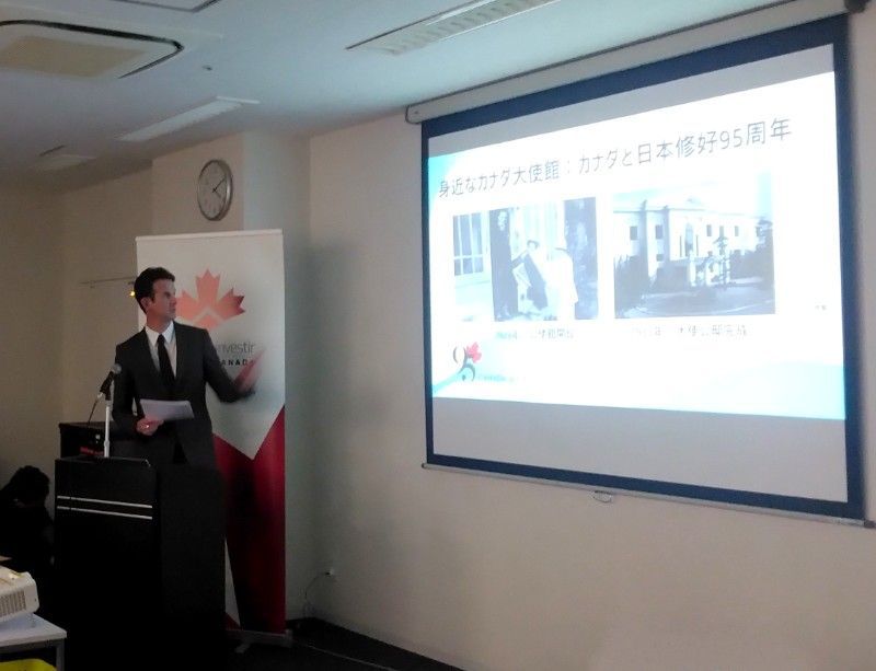 Candian Consul to Nagoya giving presentation, pointing to projected images, Canadian flag backdrop.