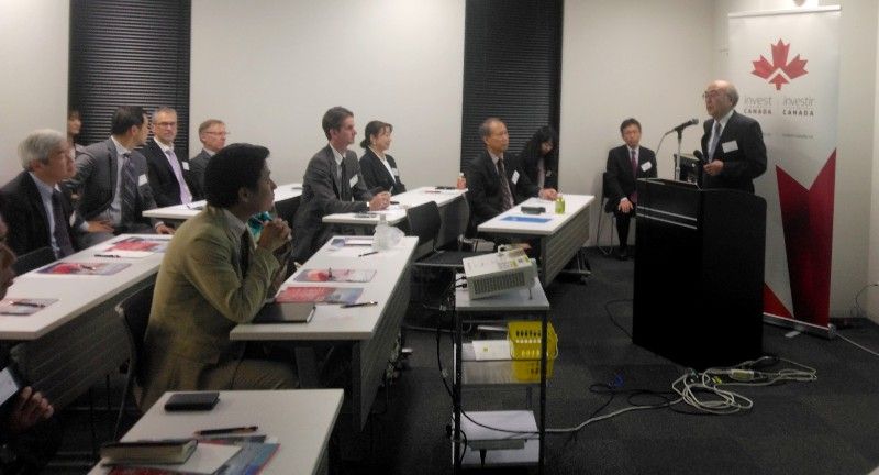 A man speaking at a podium in a conference room with attendees seated at desks. Canada logo on the banner.