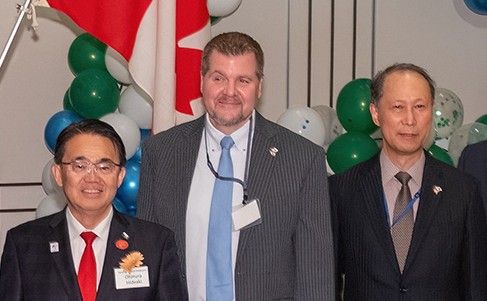 Three men in suits stand in front of a Canadian flag and balloons.