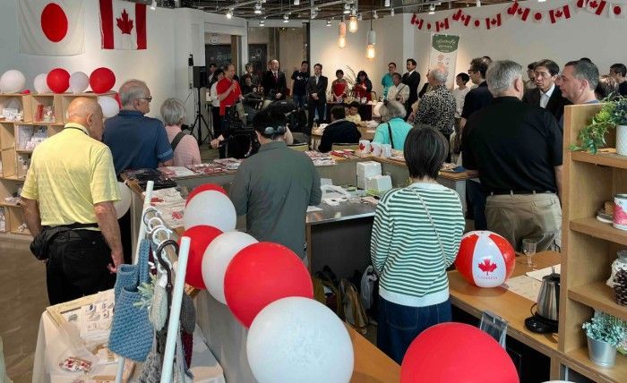 People gathered in a brightly lit room with Japanese and Canadian flags, red and white balloons, and merchandise.