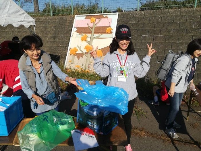 People at an outdoor event. Woman in gray hoodie giving peace sign next to trash bags on a table.