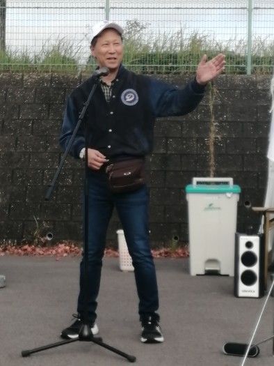 Man speaking into a microphone outdoors, gesturing with his right arm. He wears a jacket, jeans, and a white cap.