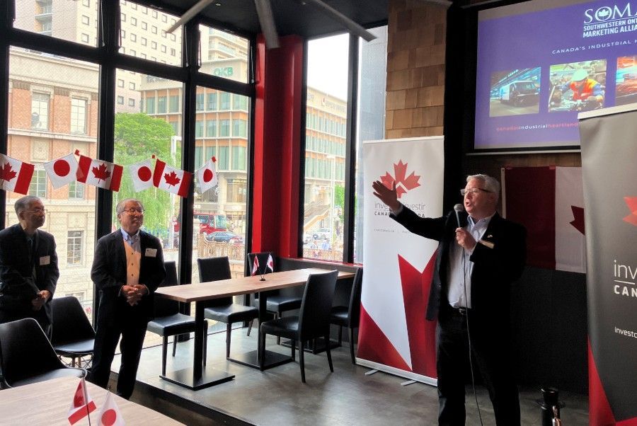 Man speaking at a Canadian event, with flags and banners. Two men listen.