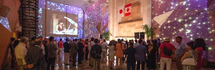 Large crowd in a room with projections and a Canadian flag. People watch a screen, some look up at aCanada Day Celebration in Nagoya.