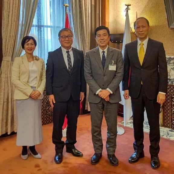 Four people, two men and two women, standing in a room with flags, a window, and a patterned wall.