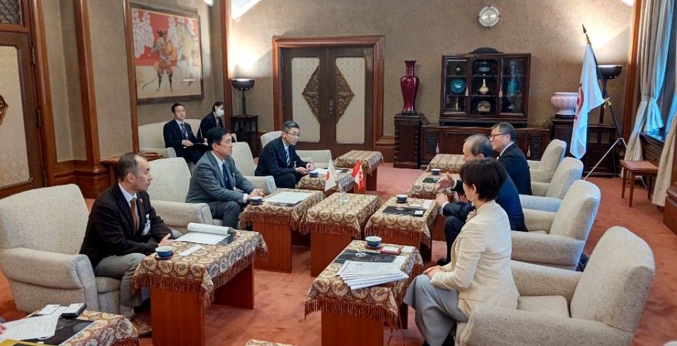 Meeting in an ornate room; multiple people seated at tables, discussing. Japanese flag present.