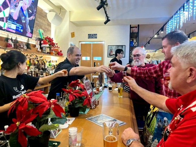 People toasting at a bar with drinks. Bartender and patrons reach hands forward. Interior with Christmas decorations.