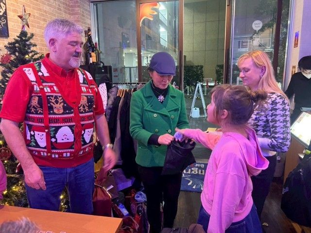 People in a shop, one giving a child a gift. A Christmas sweater, green coat, and pink shirt are visible.