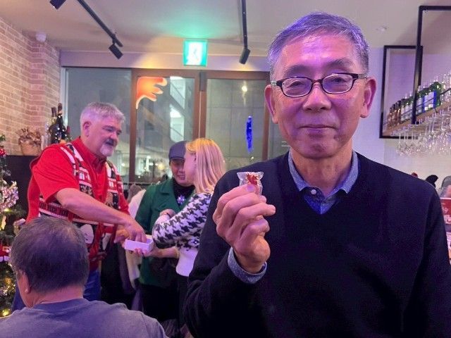 Man holding shot glass, smiling. Restaurant interior, people in background, one wearing a scarf.