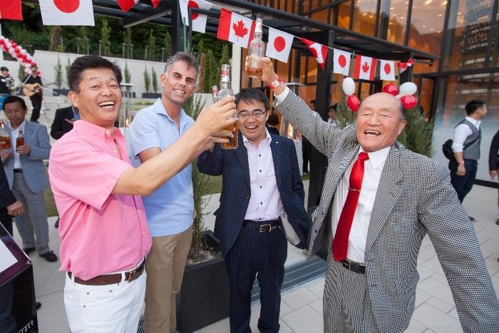 Four men toasting with bottles outdoors, Canadian flags overhead.