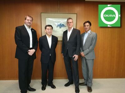 Four men in suits standing, posing in front of a wood-paneled wall and painting; Okayama logo in corner.