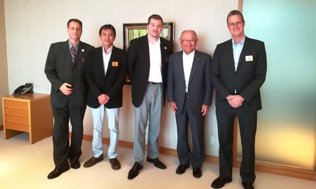 Five men in suits stand in a room with a light-colored wall, possibly a business meeting or event.