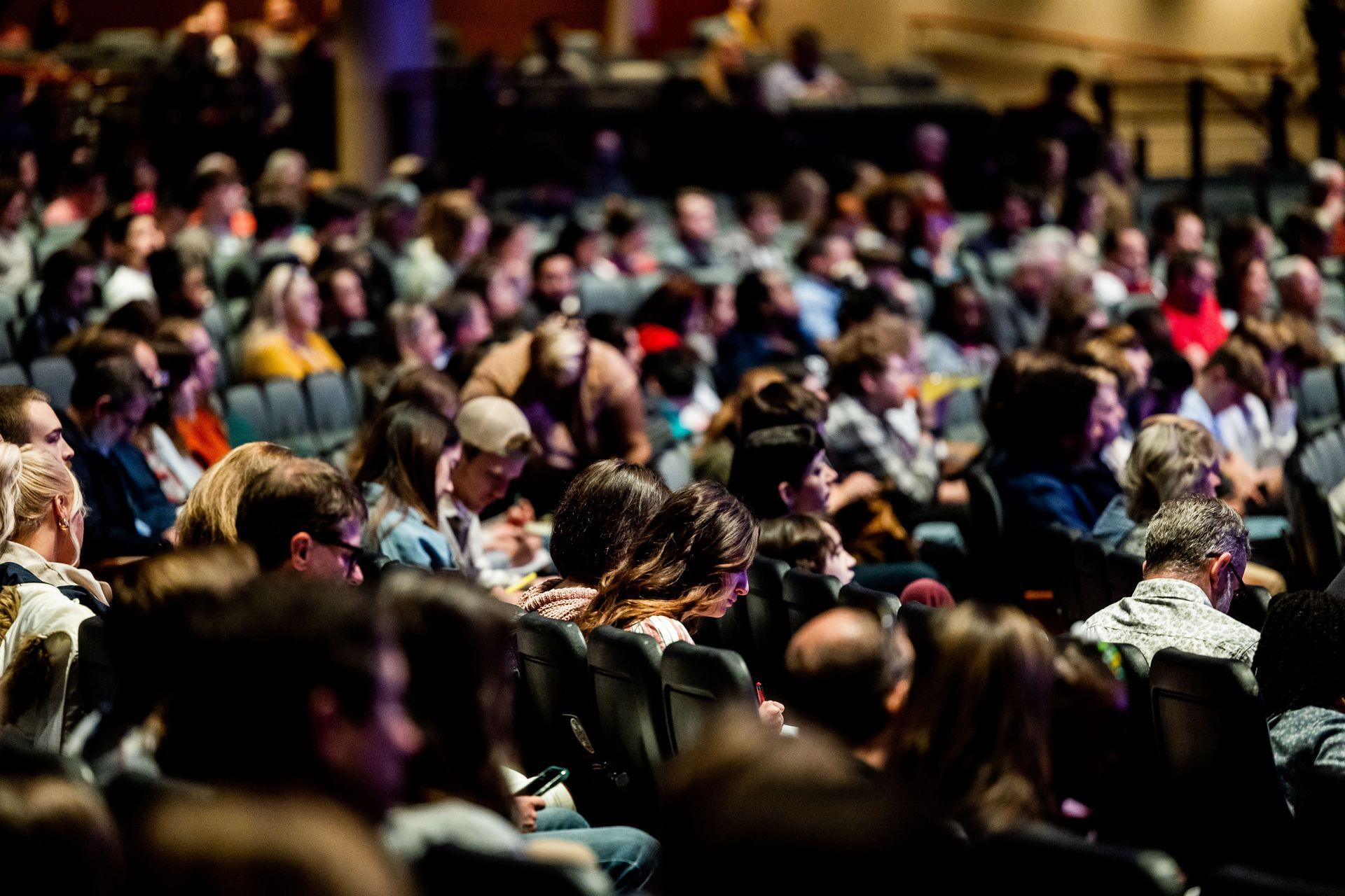 Crowded audience seated in a dimly lit auditorium, seen from behind.