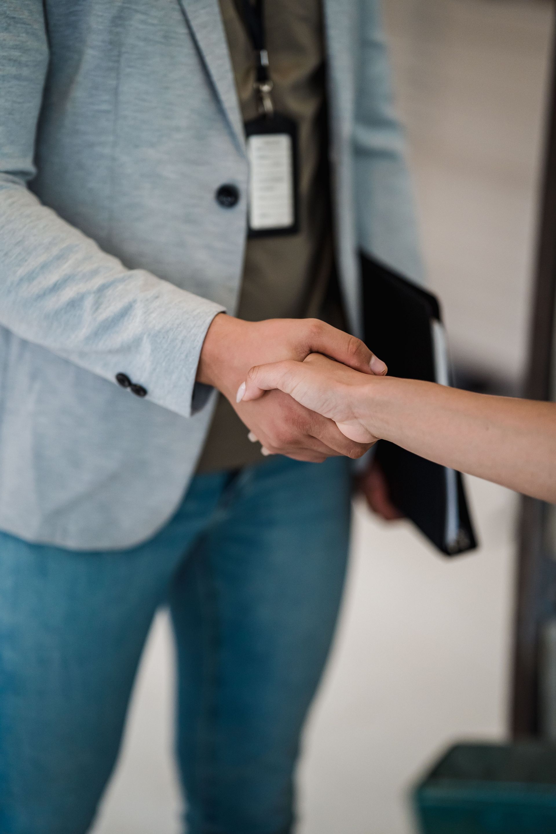 Two people shaking hands in an office, one in a light gray blazer holding a black folder.