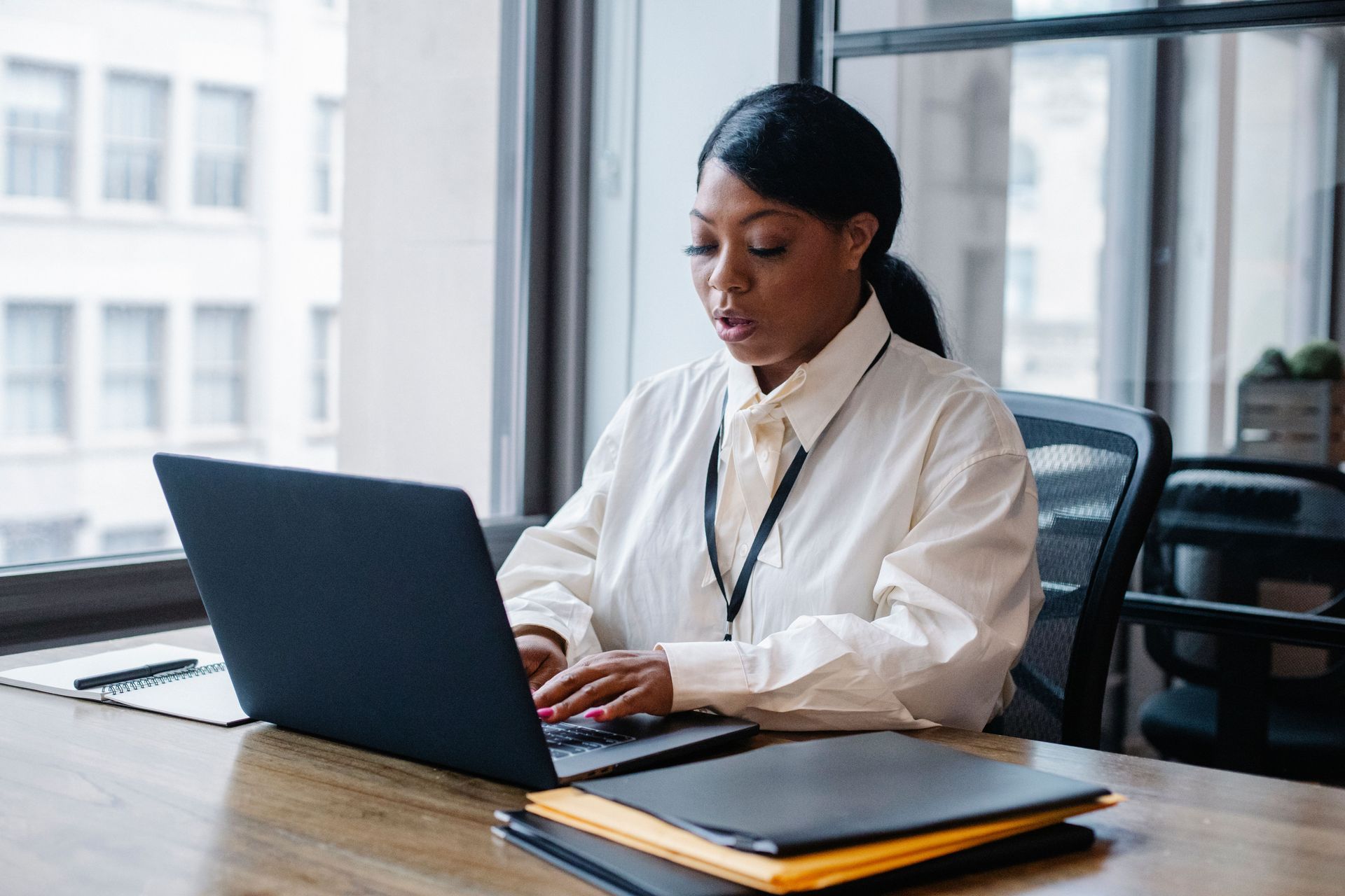 Woman working on a laptop at a desk in a bright office with a notebook and folder nearby