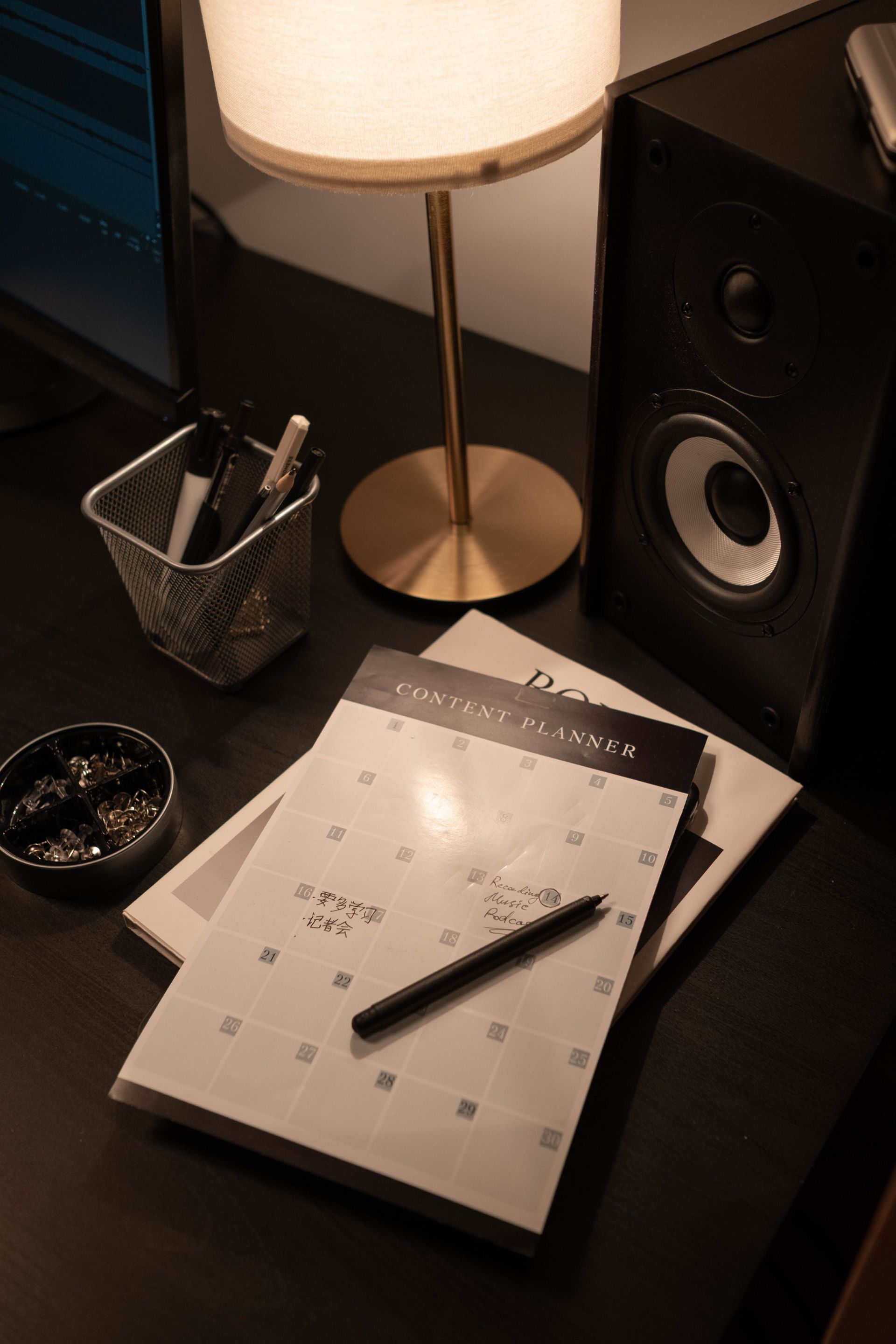 Desk with a lamp, speaker, pen holder, and a calendar notebook with a pen on top.