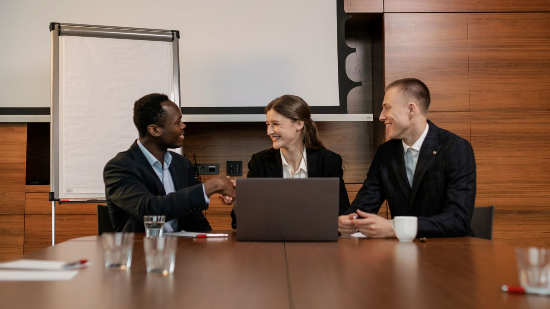 Three colleagues in a meeting, smiling and shaking hands across a conference table with a laptop and coffee mugs.