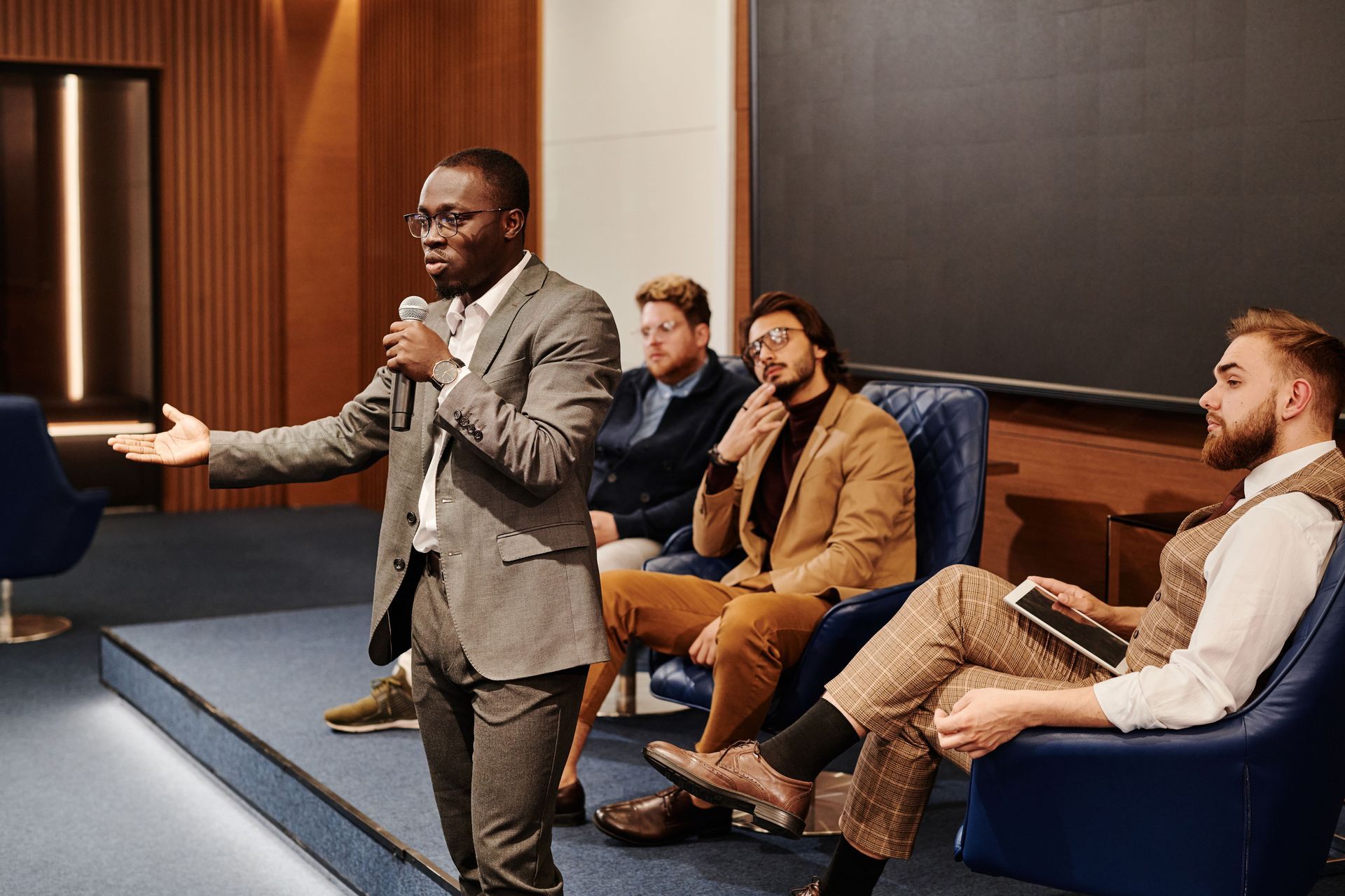 Man speaking into a microphone at a panel discussion with three seated audience members in a conference room
