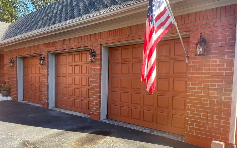 Three red-brick garage bays with matching wooden panel doors and wall-mounted lights under a gray roof with an American flag.
