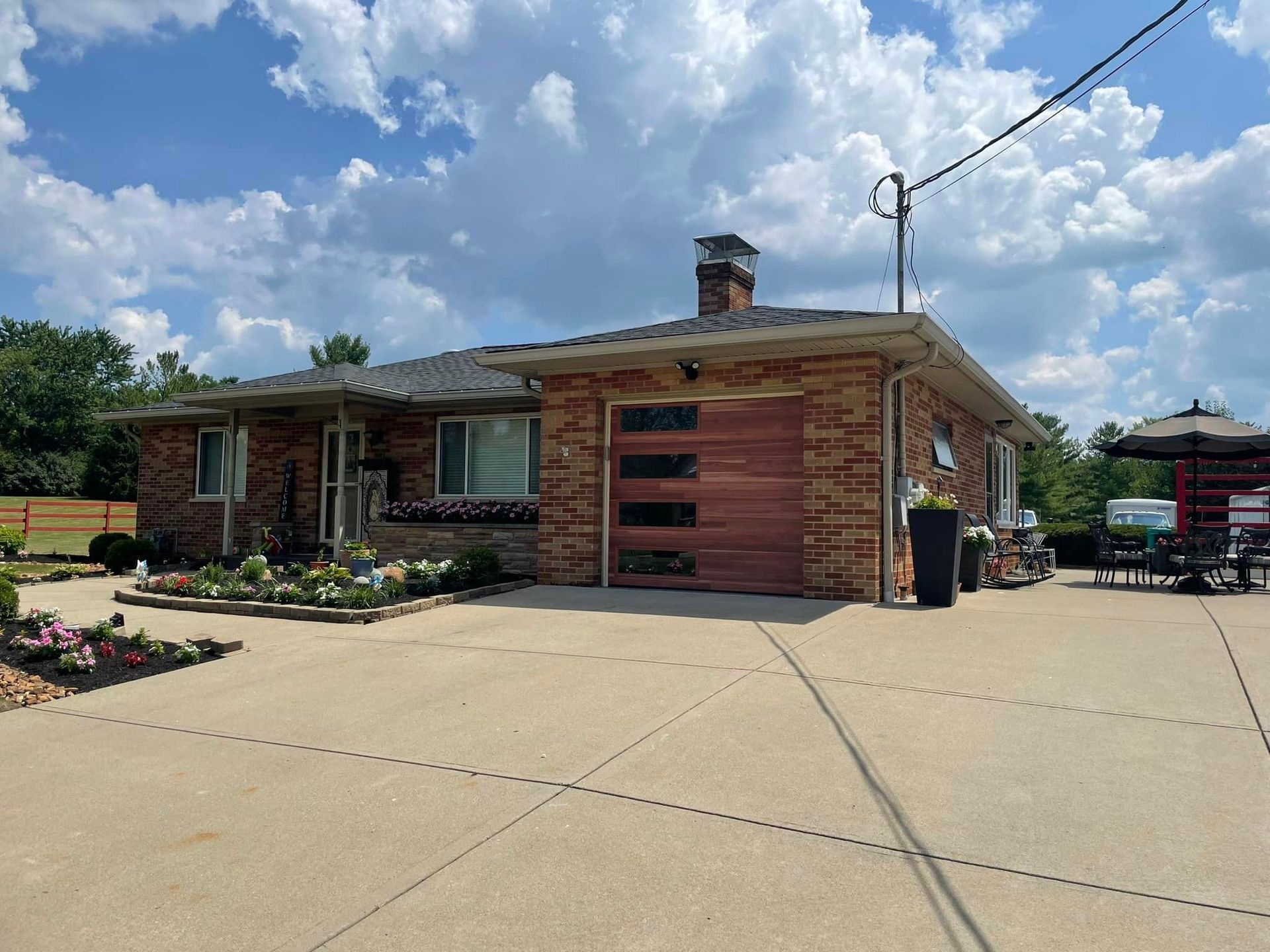 Single-story brick house with a modern wood garage door, chimney, and large concrete driveway under a cloudy sky.