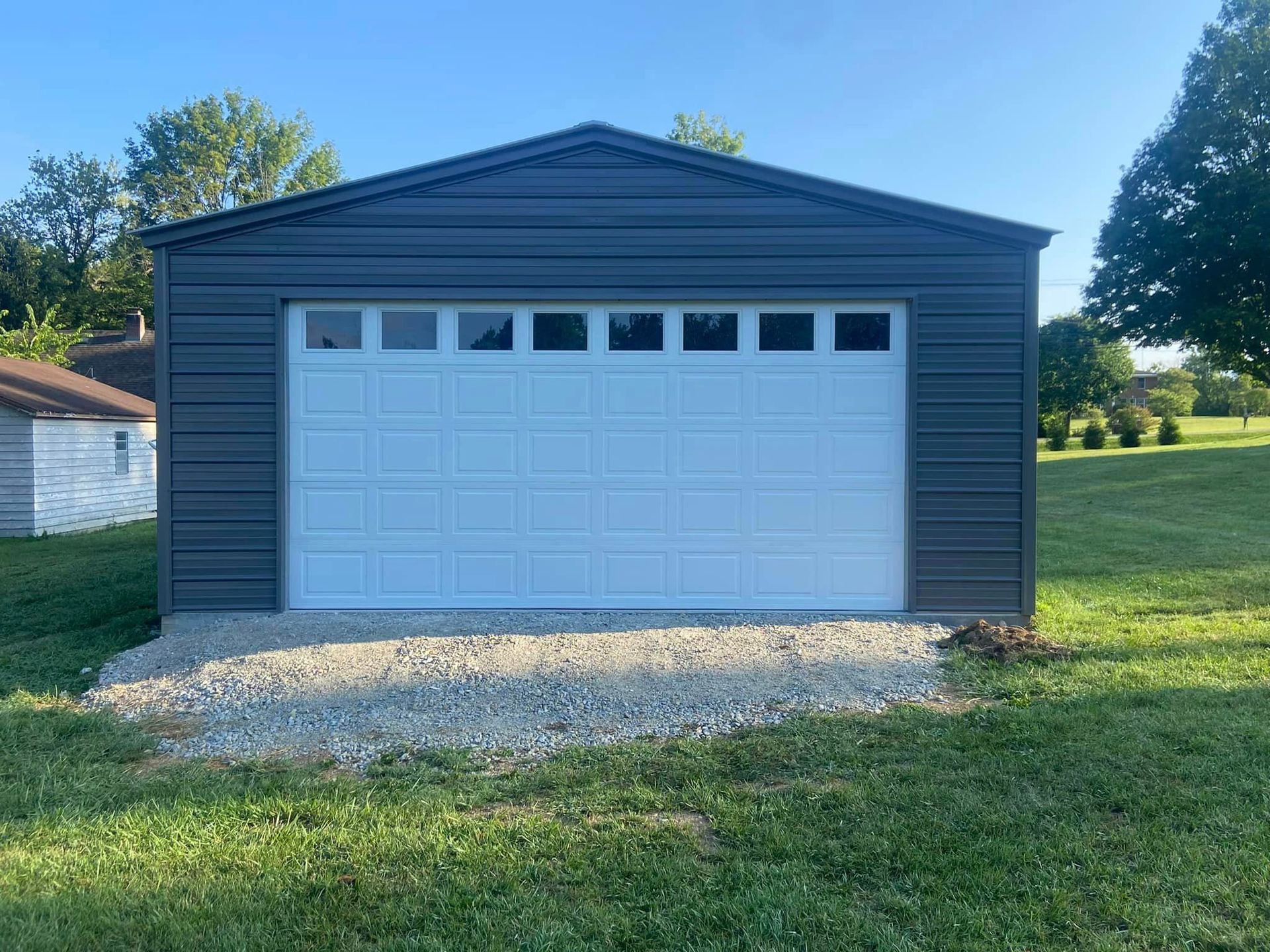 A dark gray metal garage with a large white door featuring horizontal window panels, set on a gravel pad in a grassy yard.