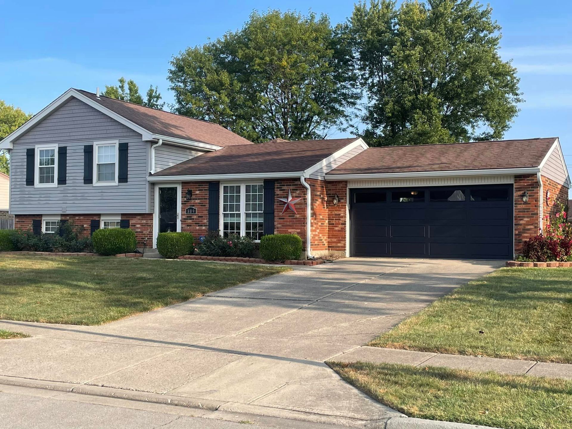A single-story, split-level house with grey siding and red brick, featuring a black garage door and a grassy front lawn.