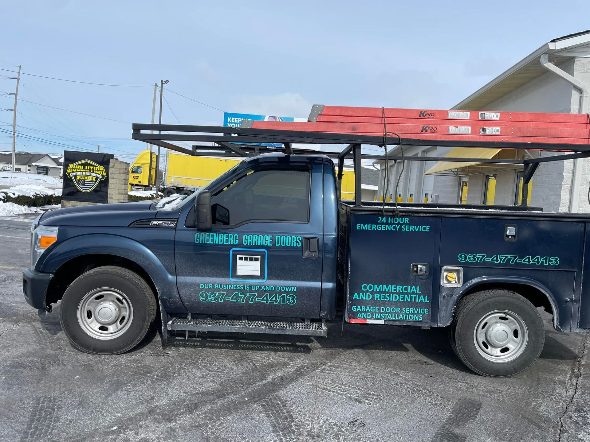 A dark blue service truck with a ladder rack and equipment decals parked outdoors on a sunny, winter day.