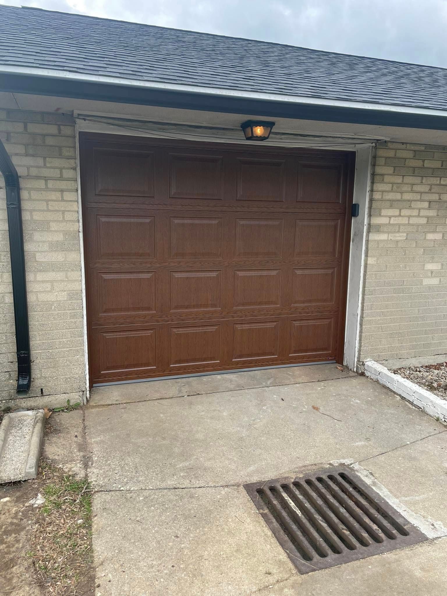 A brown garage door with rectangular panels centered on a beige brick building with a concrete driveway and drain.