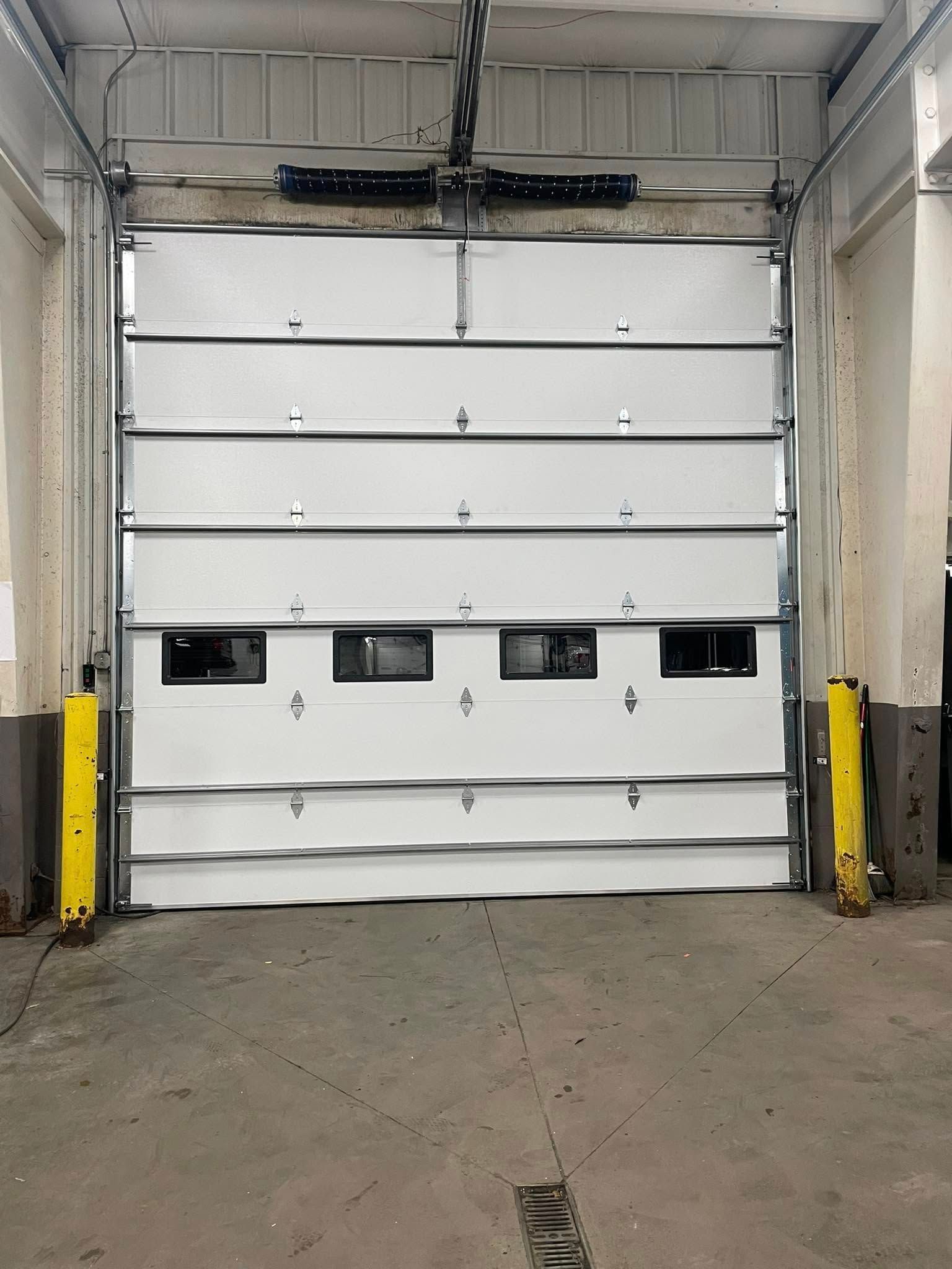 A closed, white industrial roll-up garage door with a row of small windows, framed by yellow steel bollards in a warehouse.