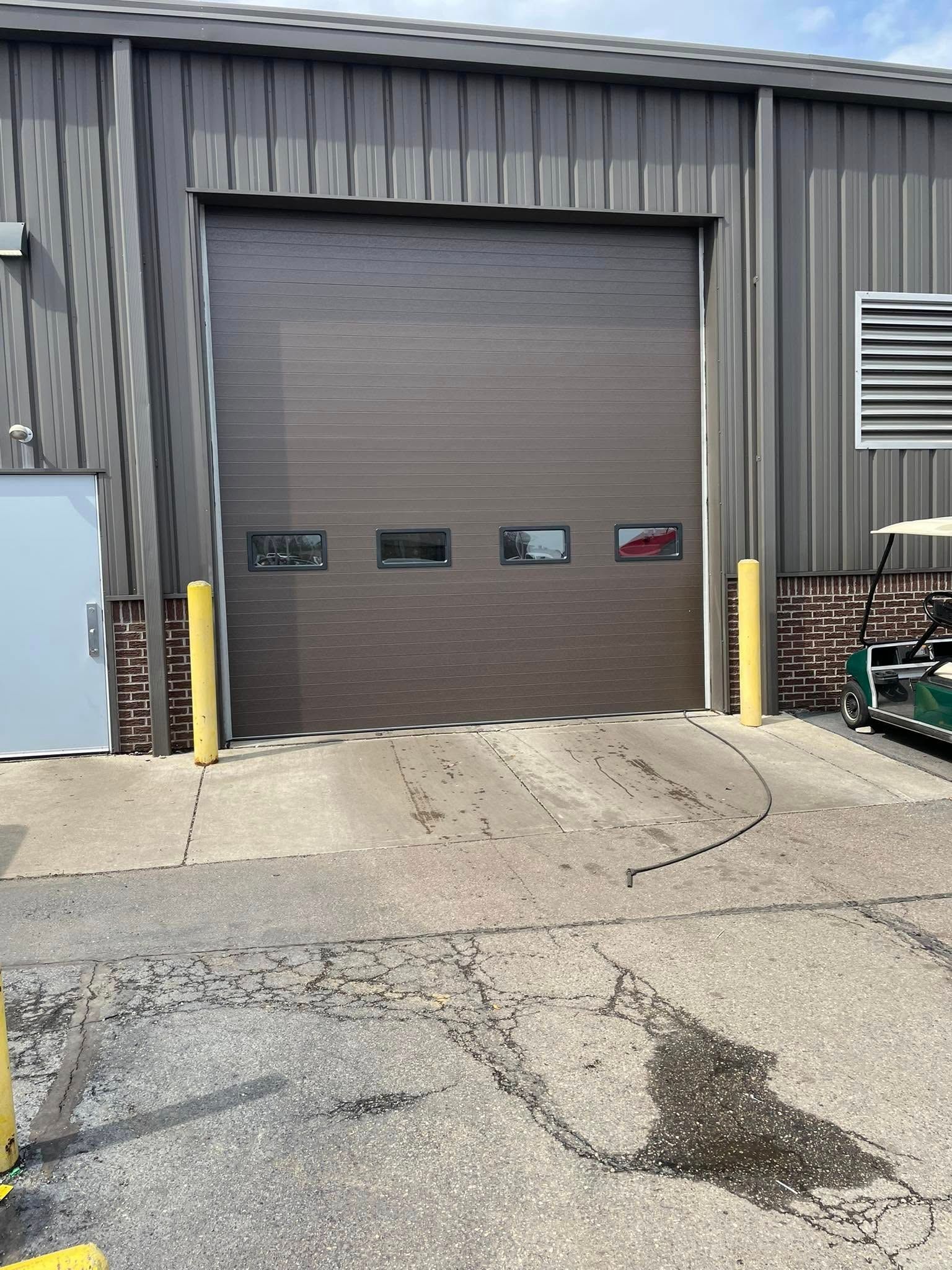 A brown, ribbed metal garage door with four small rectangular windows, installed in a grey industrial building.