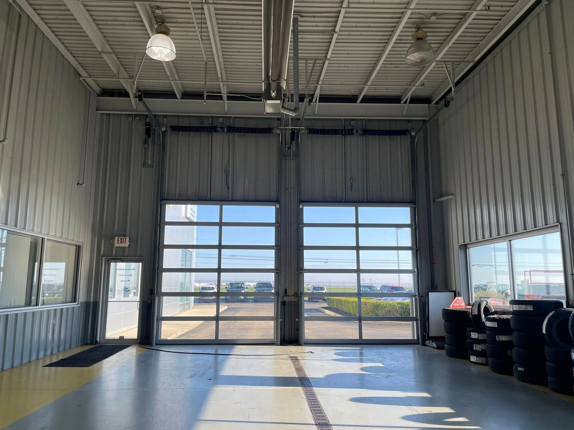 Inside of a commercial garage bay with two glass overhead doors, grey walls, and a stack of tires on the right.
