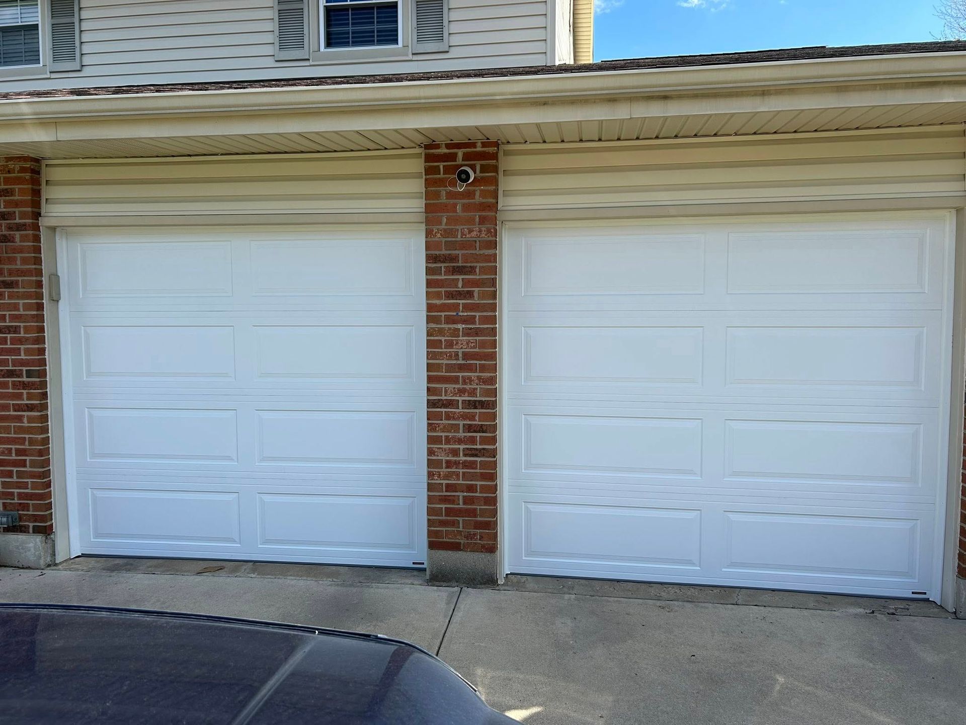 Two white garage doors with brick pillars and light-colored siding on a house exterior.