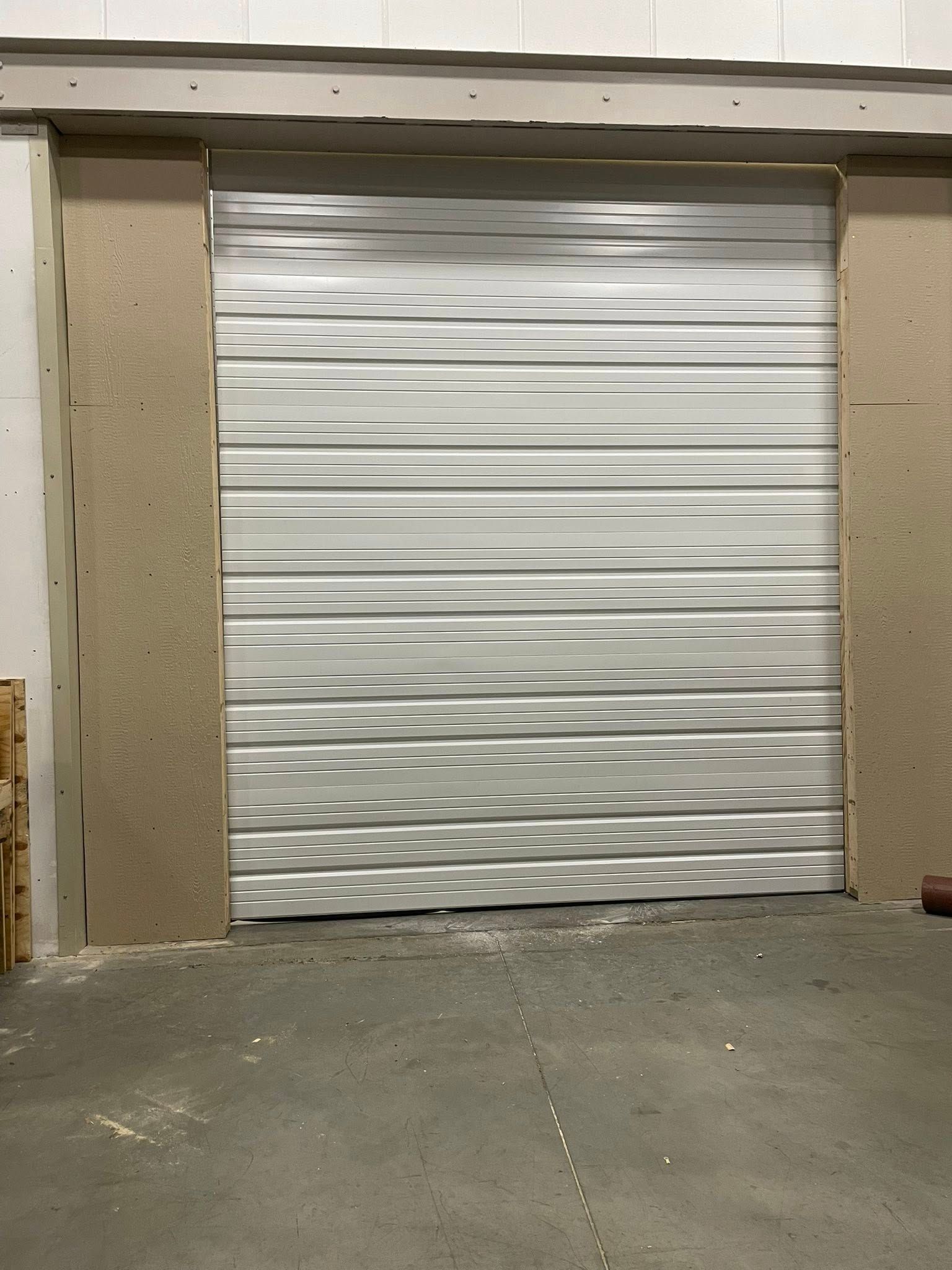 A closed silver metal rolling garage door set within a light-colored unfinished wall in a concrete-floored space.